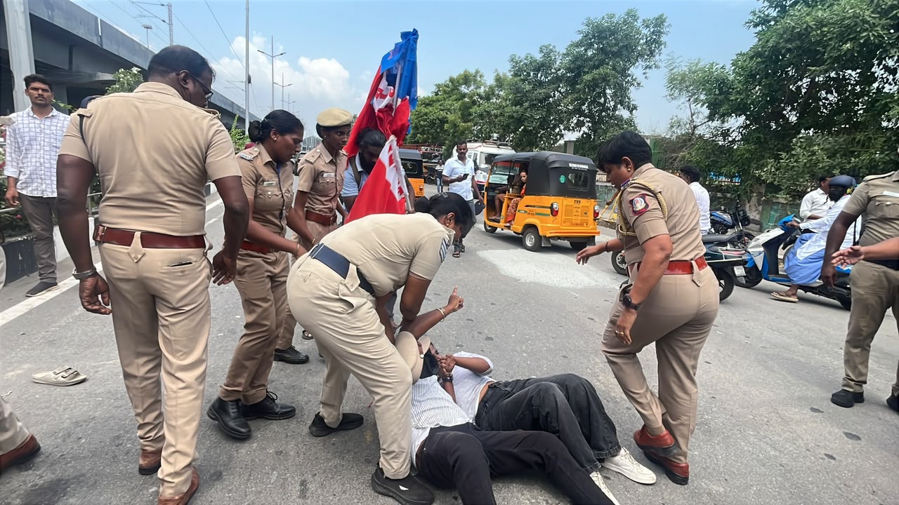 Police personnel surround and assist protesters lying on the road during a demonstration in Chennai, with AISF-AIYF flags visible in the background.