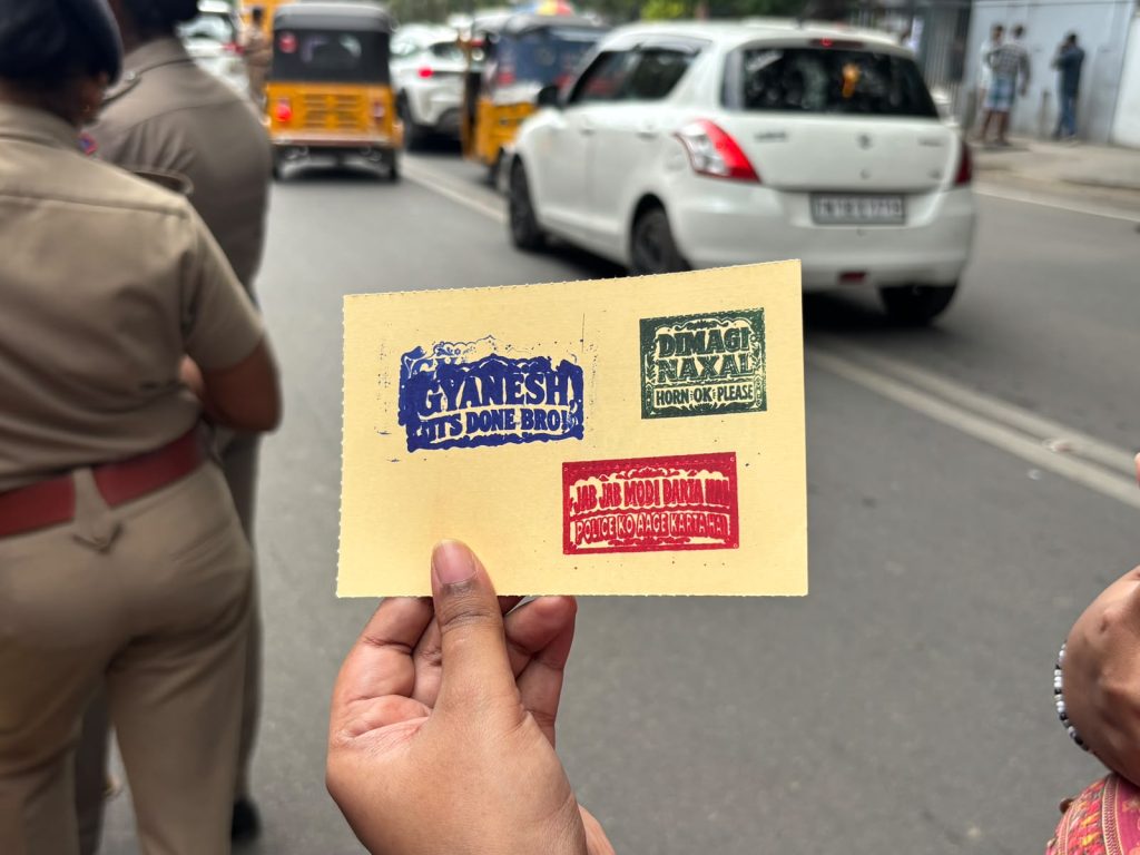 A person holds a yellow card bearing protest stickers reading “Gyanesh, it’s done bro!”, “Dimag naxal, horn OK please” and a message criticising Modi and police action, on a Chennai street.