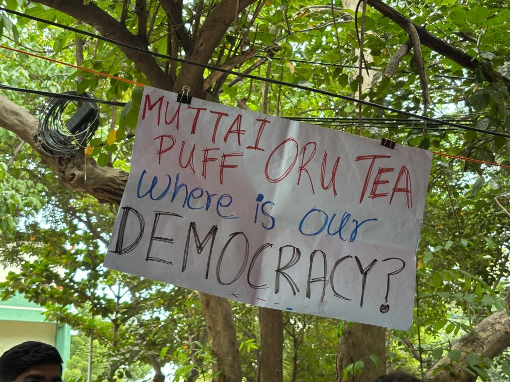 A handmade placard hanging from a tree reads, “Muttai Puff oru Tea, where is our democracy?” in red, blue and black lettering.